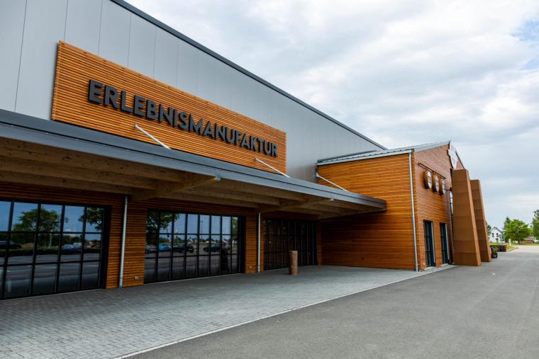 Eingangsseite mit großem Schriftzug „Erlebnismanufaktur", Holzverkleidung und auskragendem Vordach über dem gepflasterten Vorbereich