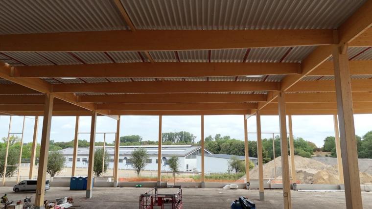 Blick aus der Halle unter dem Trapezblech-Dach auf Holzträgern nach draußen, Baustellenfahrzeuge im Vordergrund