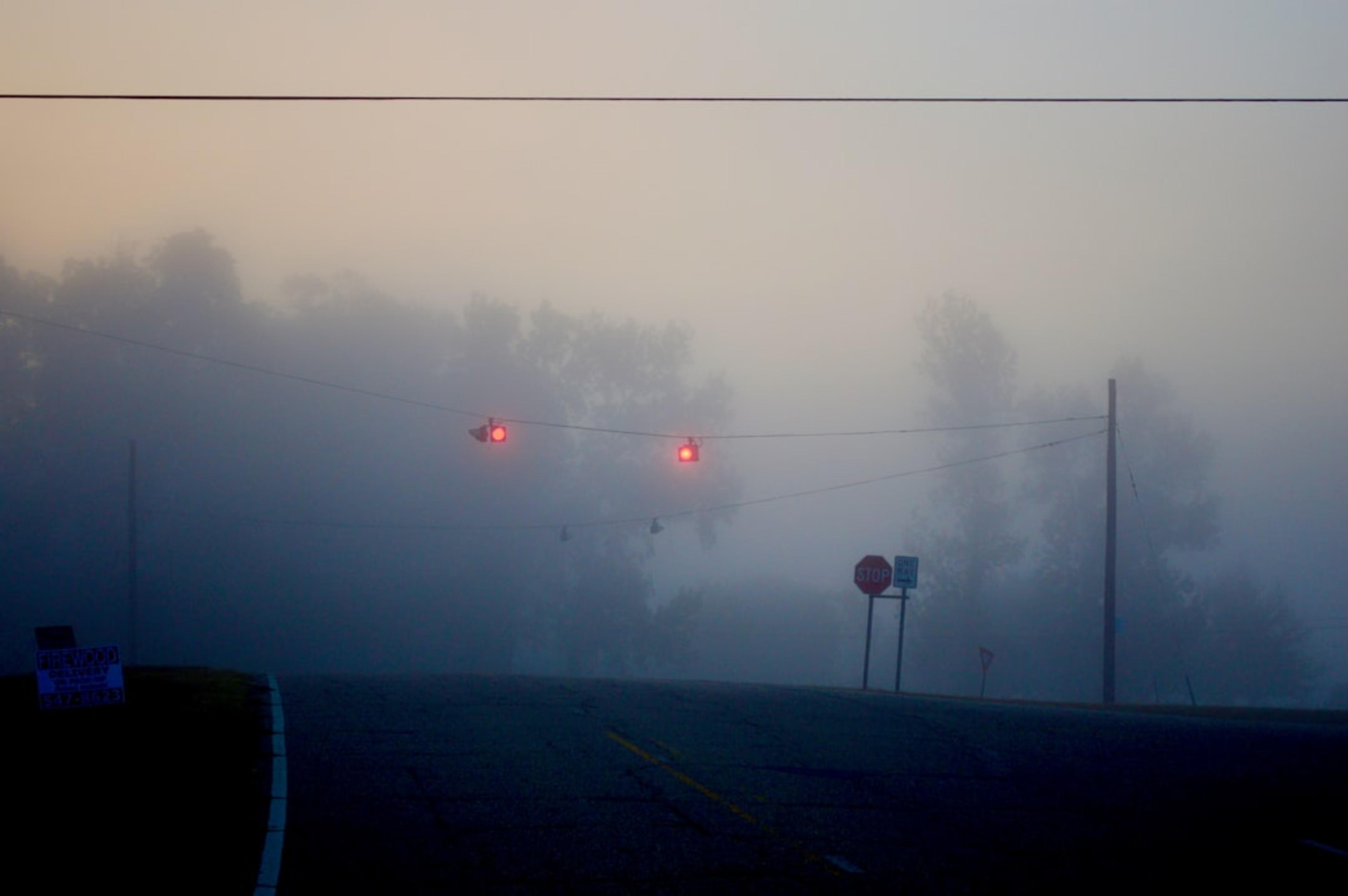 Traffic lights glow through morning fog on a road.