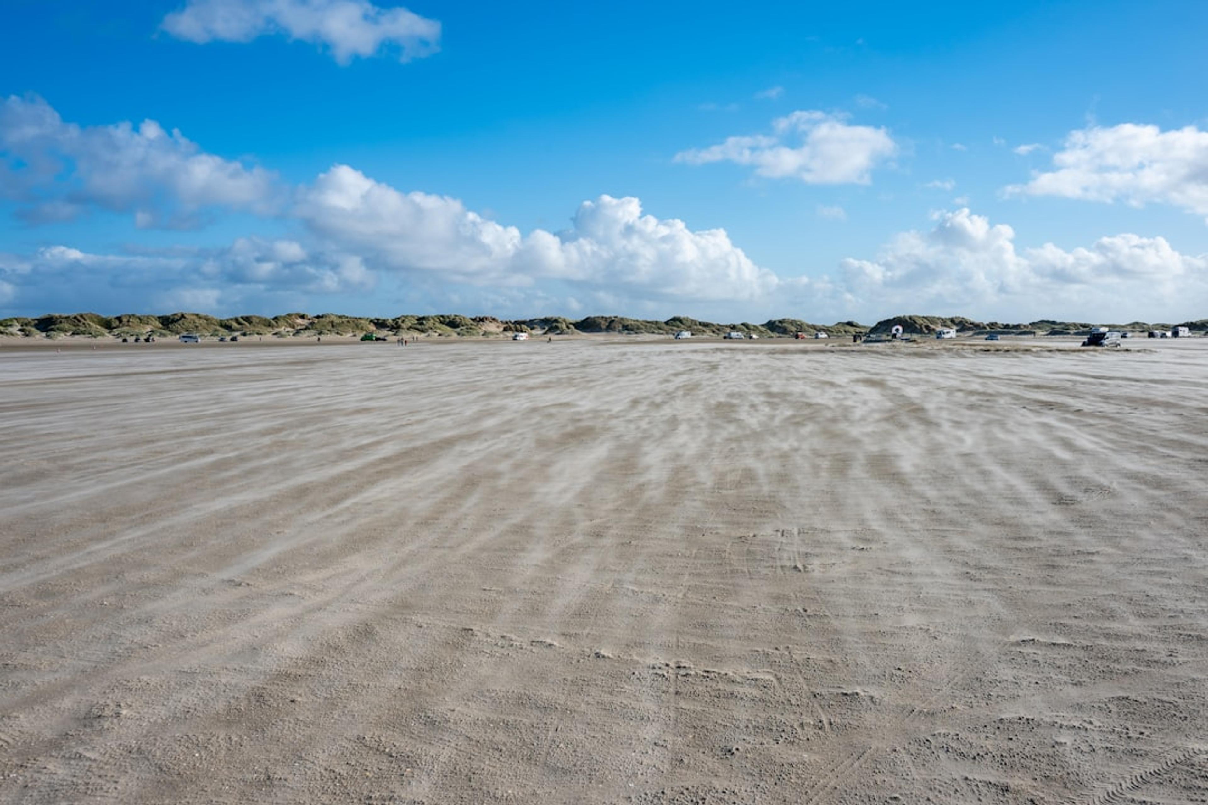 Wind blowing sand across a wide beach under blue sky.