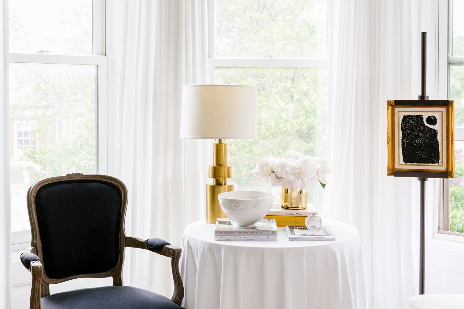 Bright transitional bedroom with black chair, gold lamp, and sheer white curtains framing garden views