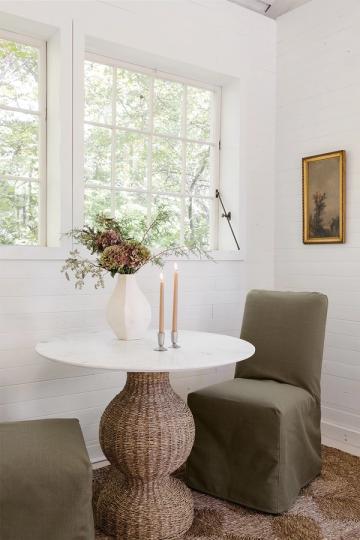 Warm minimalist dining area with white round table, woven base, and sage green slip-covered chair