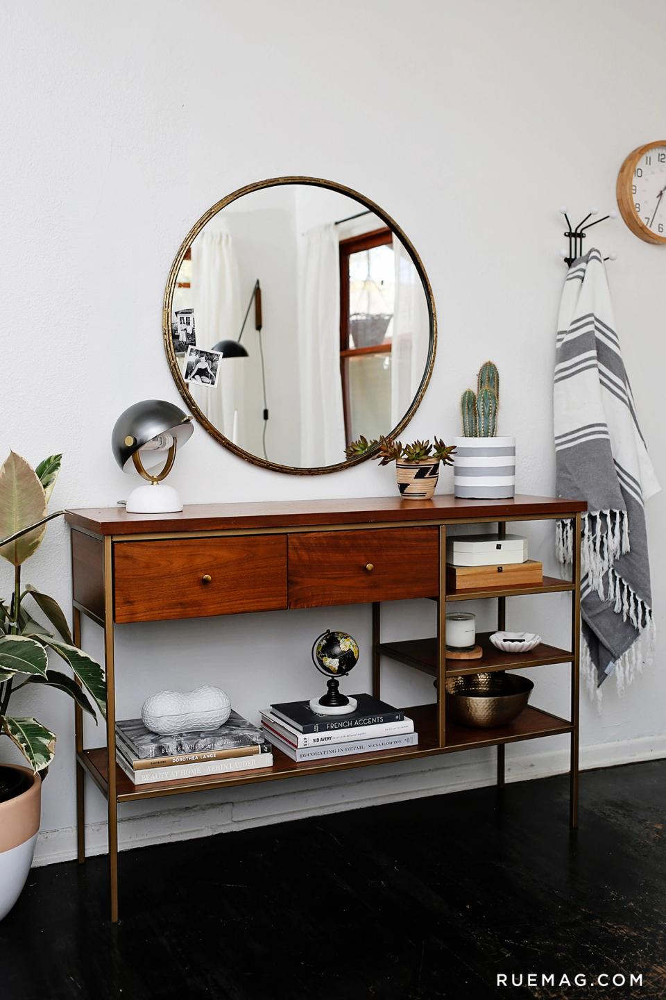 Mid-century modern entryway console with round brass mirror, wood drawers, open shelving, and minimalist plant styling