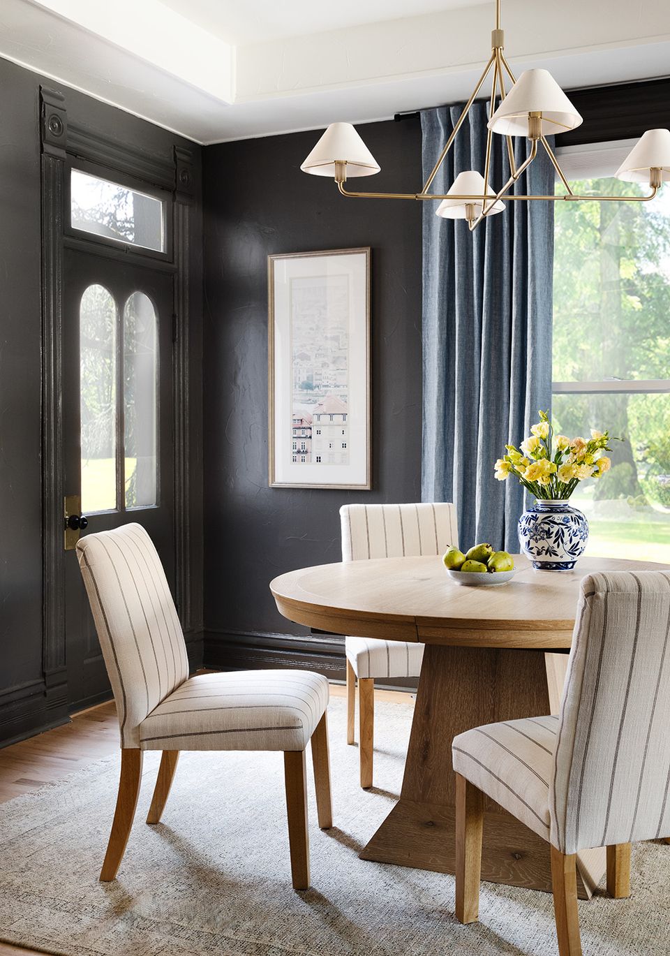 Contemporary dining room with black walls, wood round table, brass chandelier, and upholstered chairs