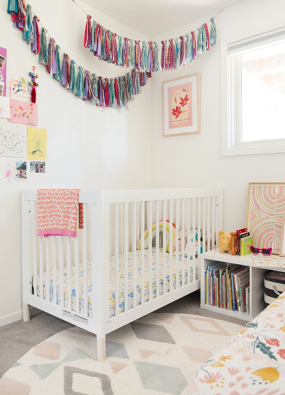 Playful modern nursery with multicolor tassel garland, pink accents, white crib, and eclectic art