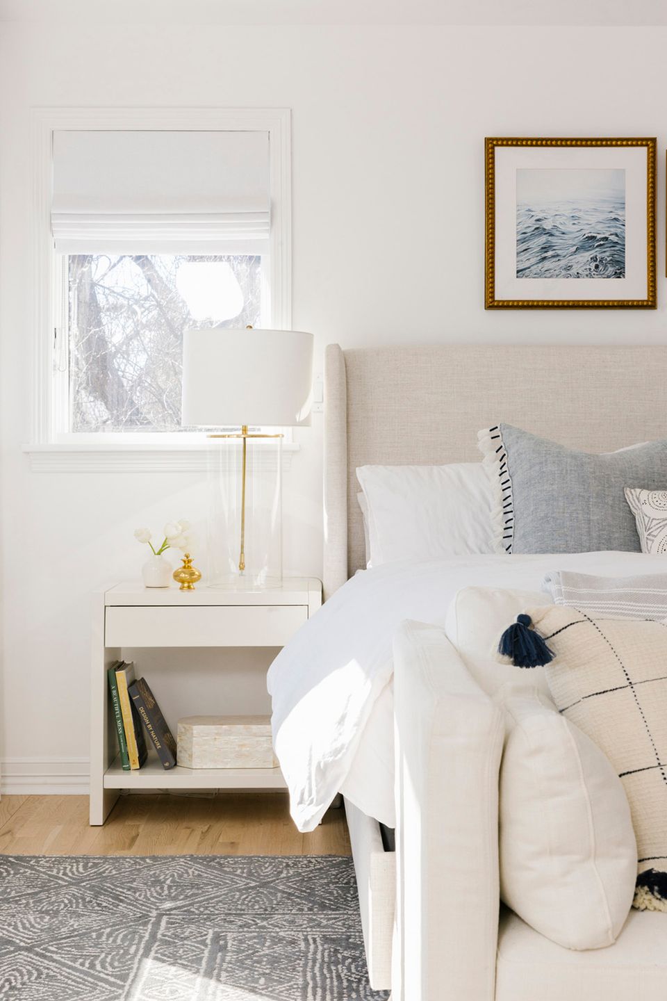 Coastal bedroom with soft upholstered headboard, brass lamp, and serene beach artwork