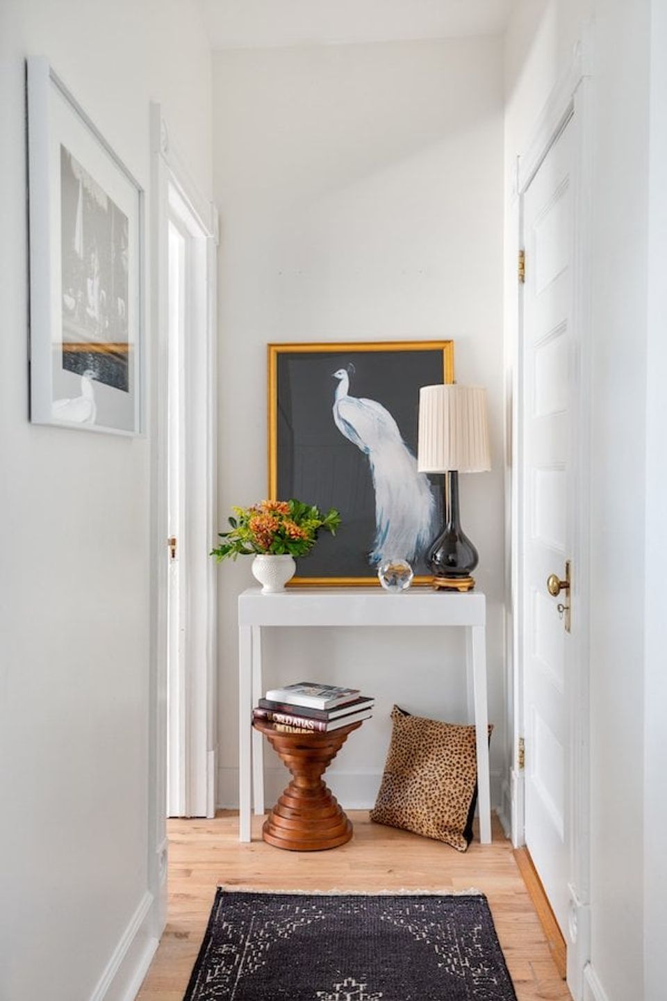 Bright minimalist hallway with white walls, wooden console table, peacock artwork in gold frame, and warm brass accents