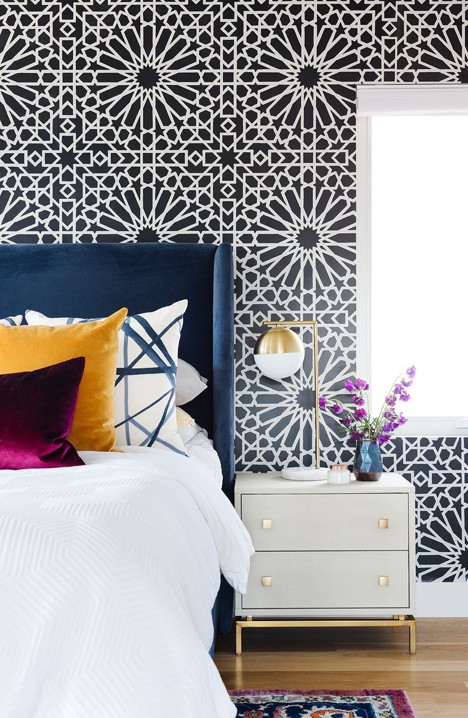 Bold eclectic bedroom featuring geometric patterned wallpaper, navy headboard, mustard and magenta accent pillows for vibrant modern style.