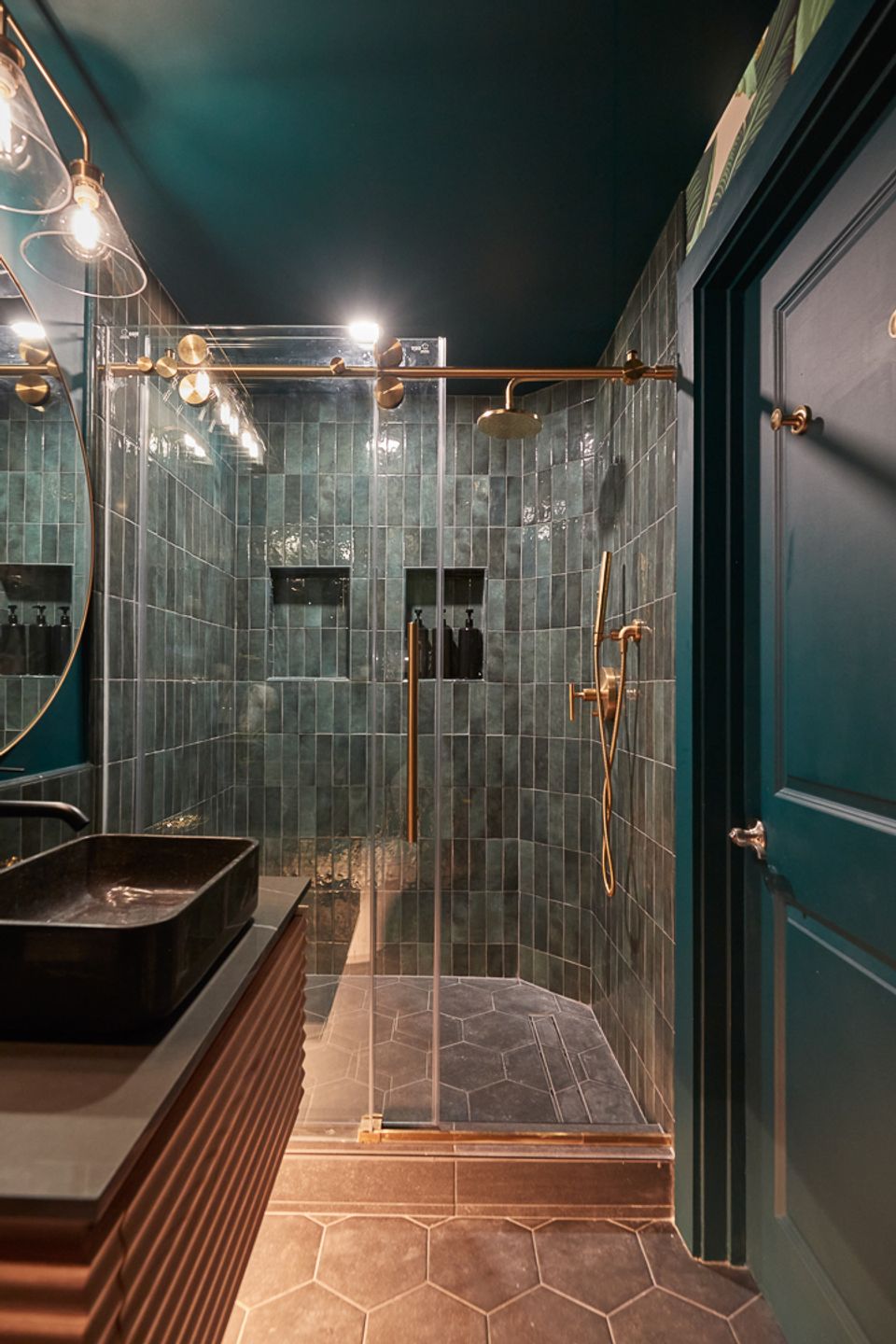 Moody emerald bathroom with dark green tile, copper fixtures, glass shower, and warm lighting