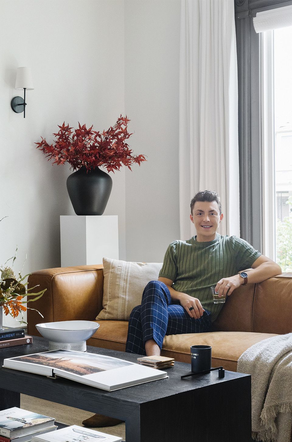 Sophisticated living space with cognac leather sofa, natural light, deep red botanical accent, and calm neutral palette.
