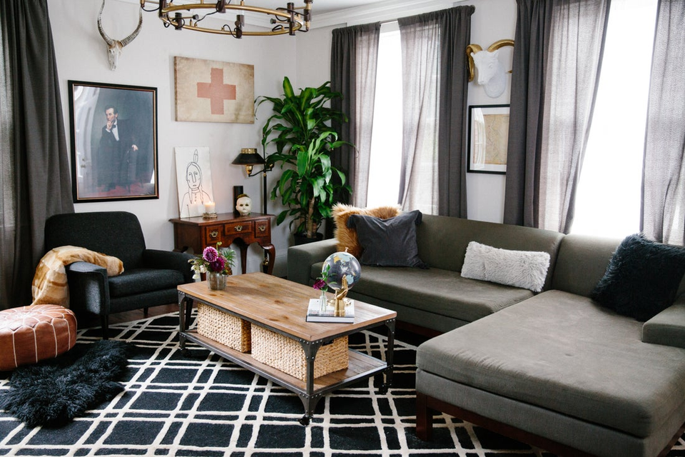 Mid-century modern living room with gray sectional, warm wood table, black geometric rug, vintage art, and lush plants.