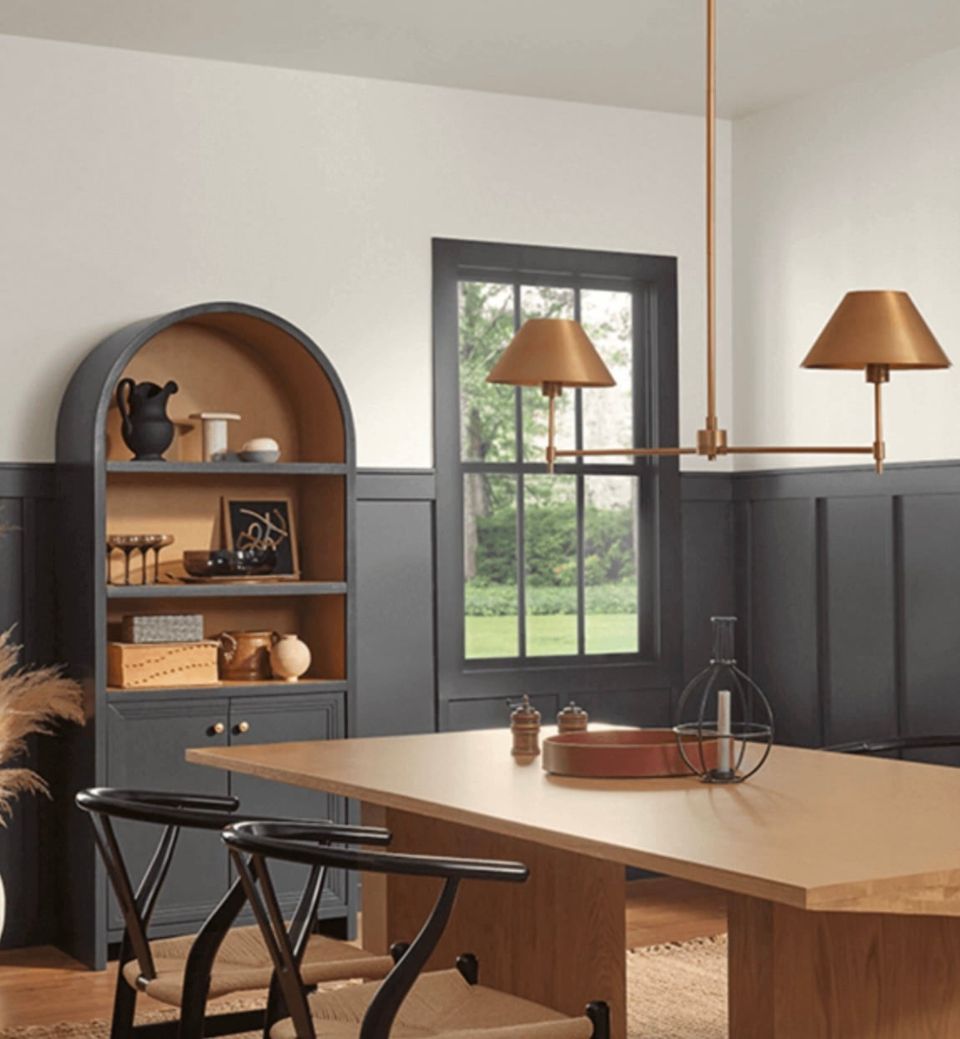 Transitional dining room with charcoal gray wainscoting, warm wood table, brass pendant lights, and arched shelving creating an inviting, layered ambiance