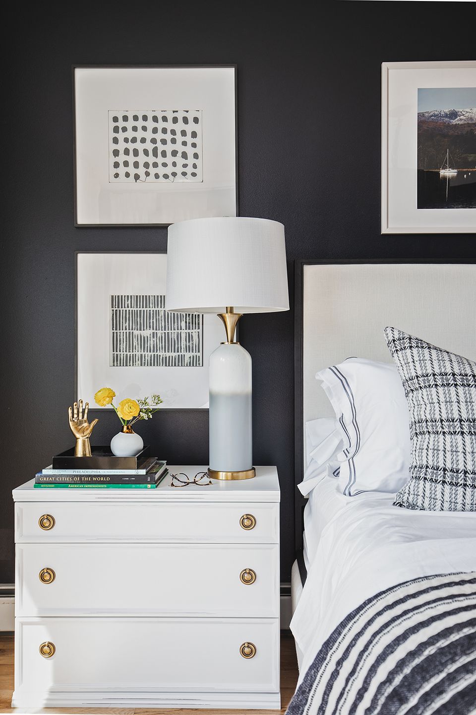 Modern eclectic bedroom with dark charcoal accent wall, white nightstand with brass hardware, and framed abstract art in sophisticated black and white tones.
