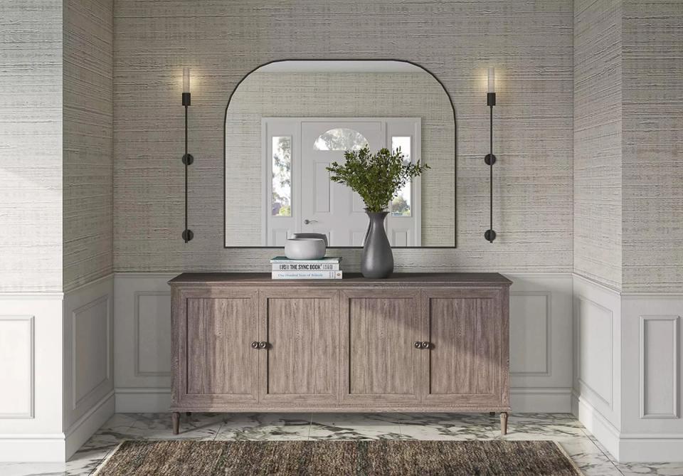 Transitional bathroom vanity with arched mirror, sconce lighting, and warm wood cabinetry on neutral tile walls.