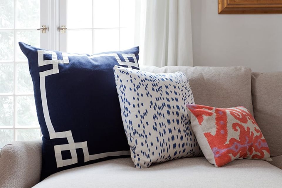 Contemporary college dorm with geometric patterned pillows in navy, white, and coral on a neutral upholstered bed by bright windows