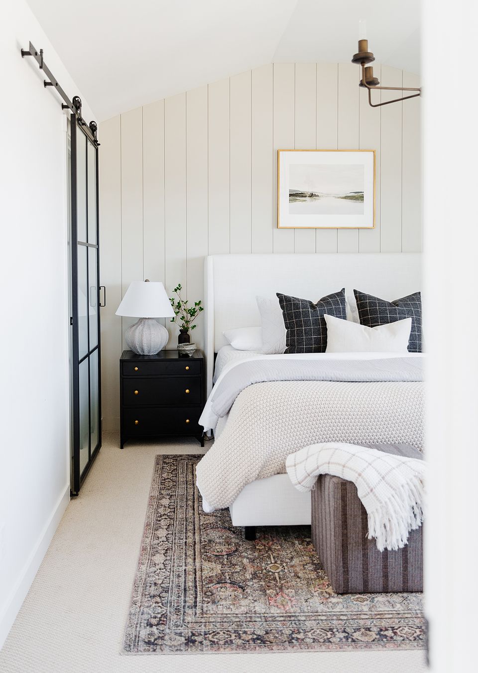Serene farmhouse guest bedroom with shiplap walls, black metal bed frame, checkered pillows, and neutral layered textures.