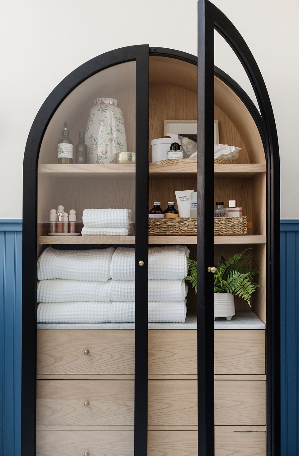 Coastal-inspired storage cabinet with arched black frame, natural wood shelving, blue wainscoting, and organic display of linens and greenery.
