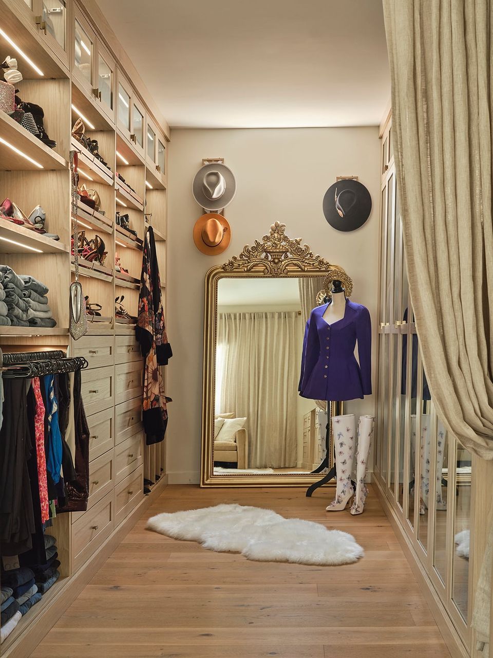 Glamorous walk-in closet with built-in shelving, ornate gold mirror, cream curtains, and curated fashion display in neutral tones.