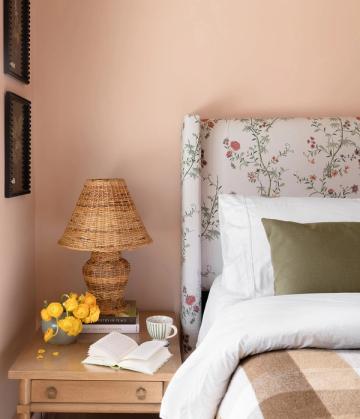 Warm, traditional bedroom with blush pink walls, floral upholstered headboard, woven rattan lamp, and layered neutral bedding with olive accent pillow