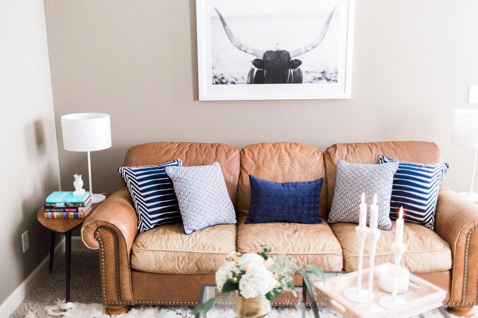 Warm transitional living room with caramel leather sofa, navy and striped pillows, neutral walls, and modern artwork