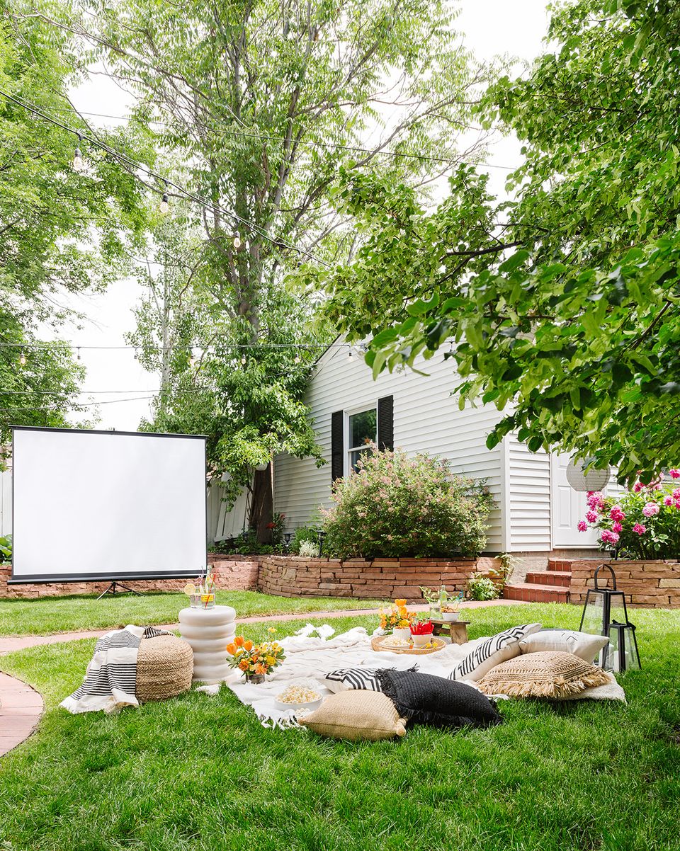 Casual outdoor movie setup on lush lawn with cozy blankets, natural wood seating, and relaxed family gathering vibe.