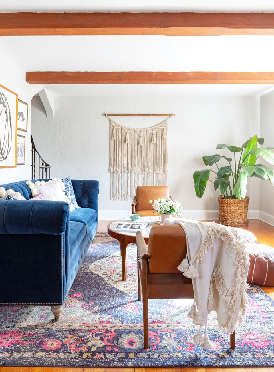 Eclectic boho living room with navy velvet seating, macramé wall hanging, warm wood beams, and vibrant layered rug