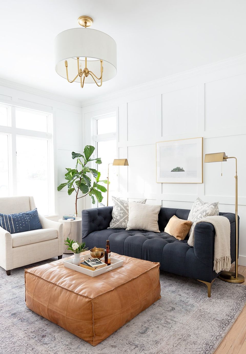 Contemporary living room with navy sectional sofa, warm leather ottoman, brass fixtures, and cream accents in bright, minimalist-modern space