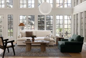 Bright minimalist living room with floor-to-ceiling windows, cream sofa, dark green chair, and natural wood accents