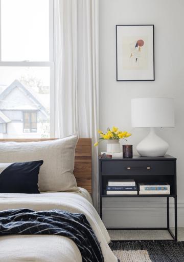 Contemporary bedroom nook with cream bedding, black nightstand, and botanical art showcasing modern simplicity with warm wood details.