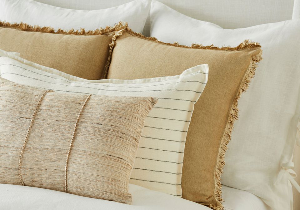 Warm neutral bed styling with layered tan and cream textured pillows and frayed edges for cozy seasonal appeal