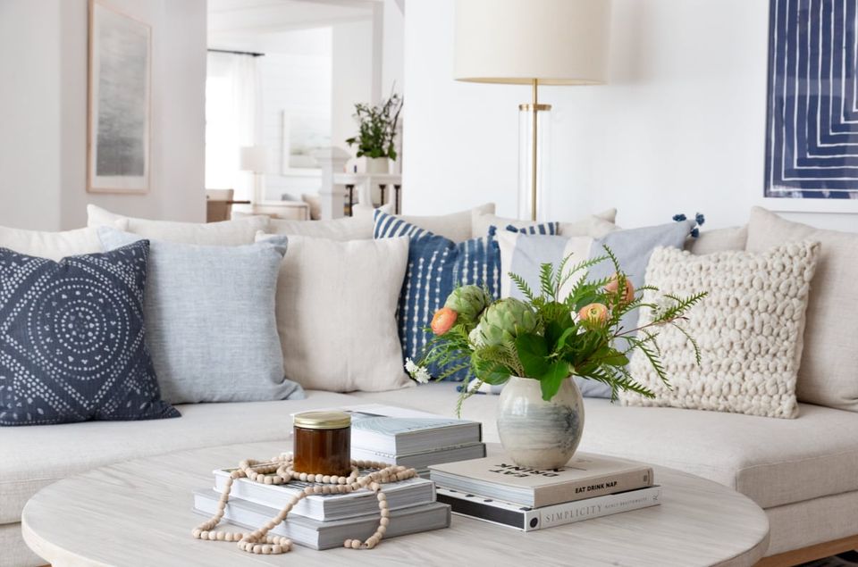Coastal living room with cream sofa, navy patterned pillows, white round table, and fresh greenery styling in soft, serene tones