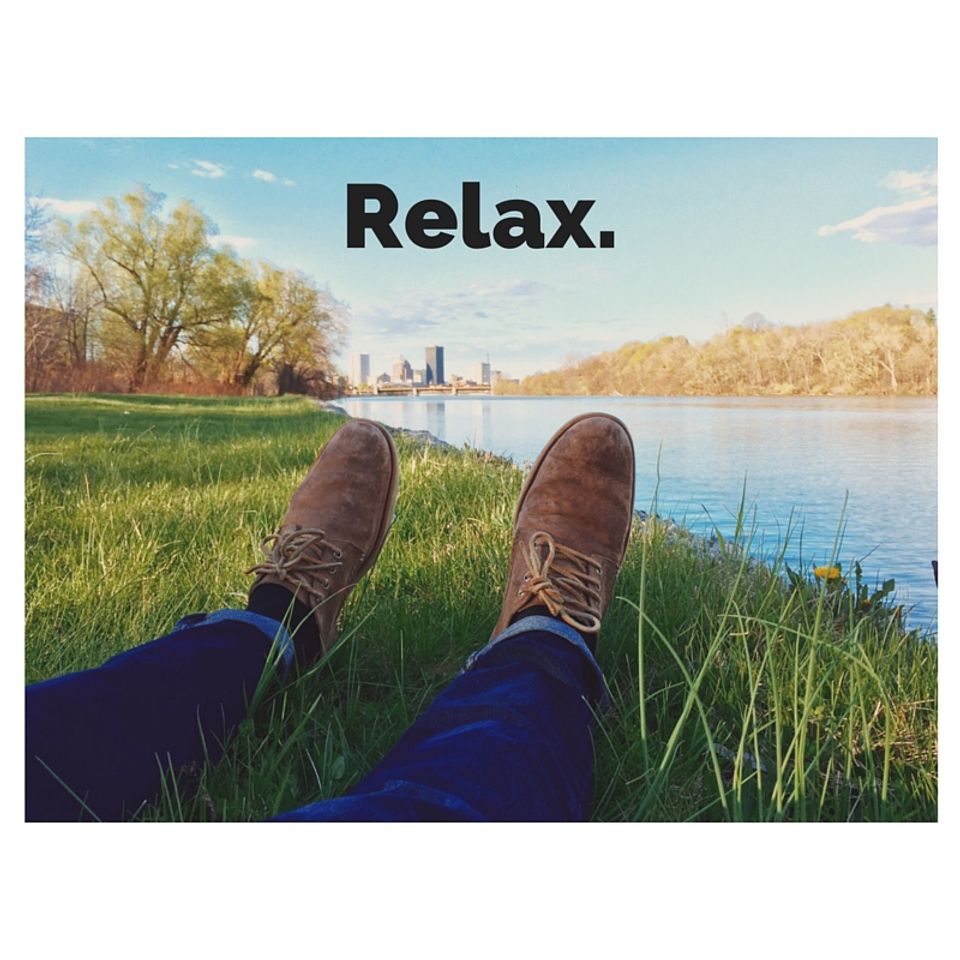 Serene waterside relaxation scene with boots propped on grass overlooking calm river and distant skyline