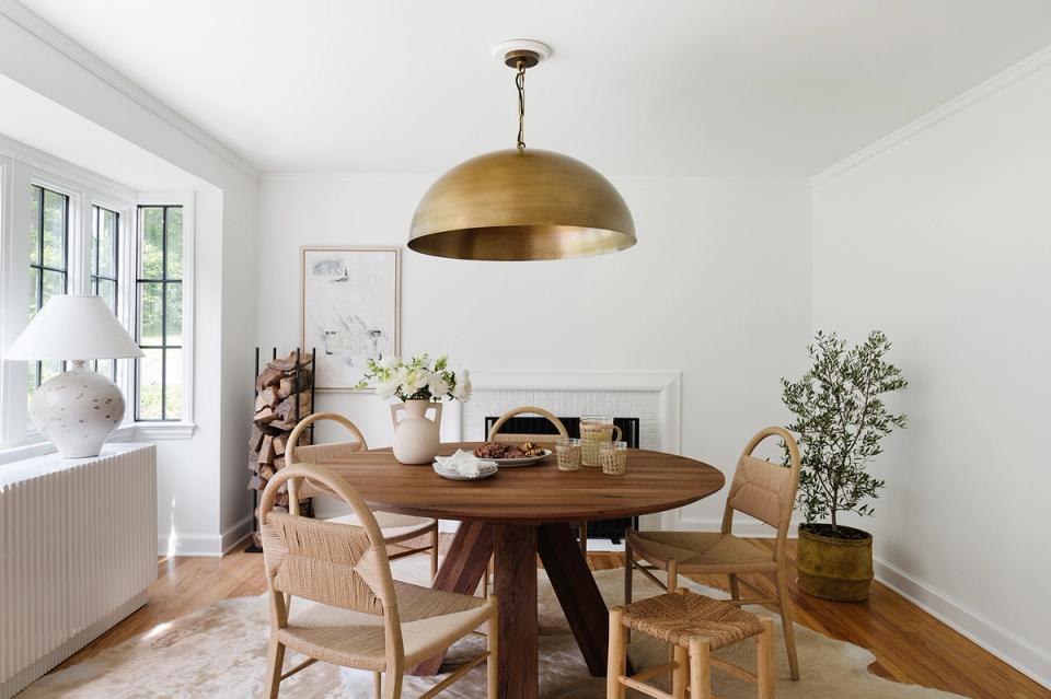 Bright, minimalist dining space with mid-century modern aesthetic featuring warm wood round table, natural rattan chairs, and brass pendant light above.