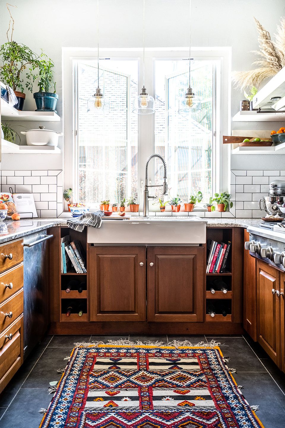 Eclectic kitchen mixing warm wood cabinetry, white subway tile, colorful layered rug, and industrial pendants