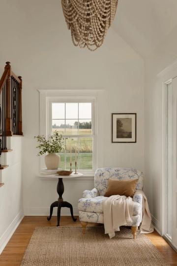 Serene farmhouse entryway with cream walls, beaded chandelier, and floral upholstered chair by window overlooking pastoral landscape