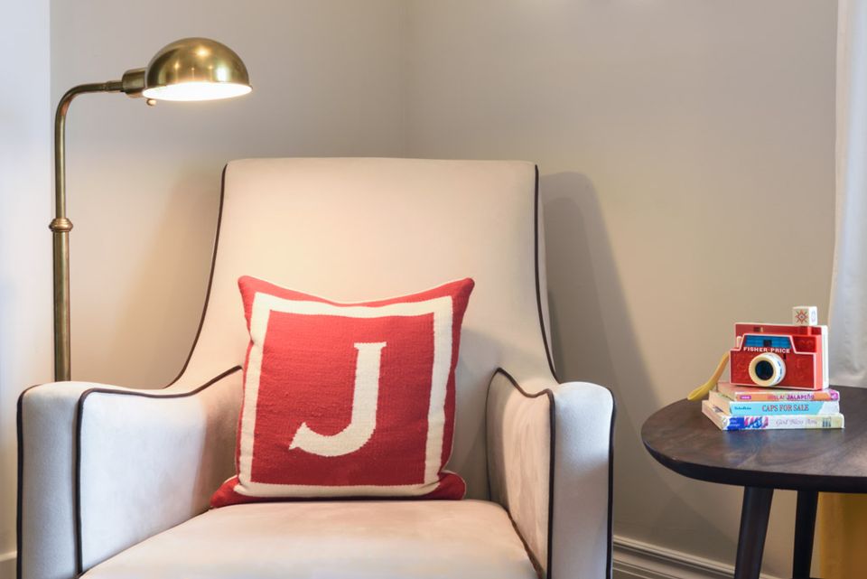 Cozy nursery corner with cream armchair, coral monogram pillow, and warm brass lighting