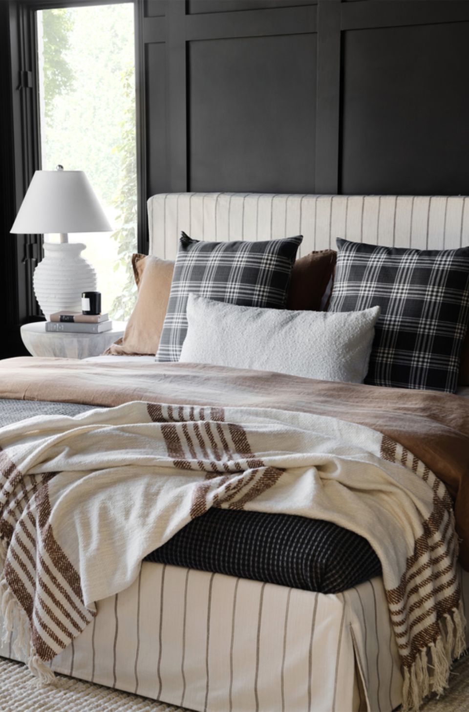 Contemporary dark bedroom with black paneled walls, cream striped headboard, plaid pillows, and neutral layered bedding