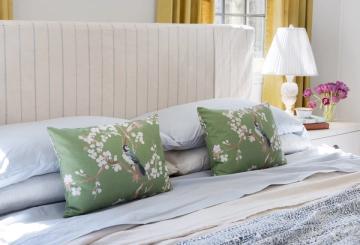 Fresh coastal-inspired bed arrangement with cream headboard, green floral pillows, and soft layered linens in whites and blues