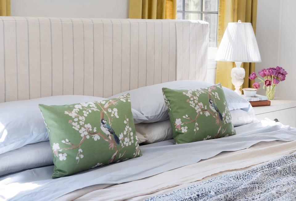 Fresh coastal-inspired bed arrangement with cream headboard, green floral pillows, and soft layered linens in whites and blues