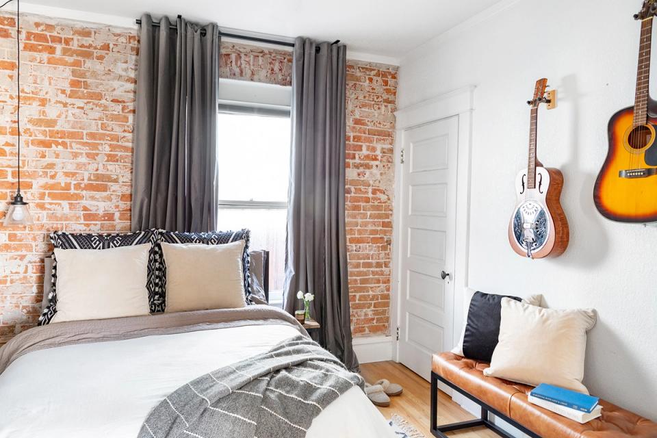 Industrial bedroom with exposed brick walls, guitars as wall art, and moody gray curtains against neutral palette