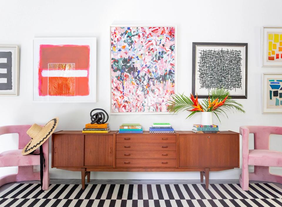 Playful maximalist workspace with pink accent chairs, mid-century credenza, colorful abstract art, and bold striped rug.
