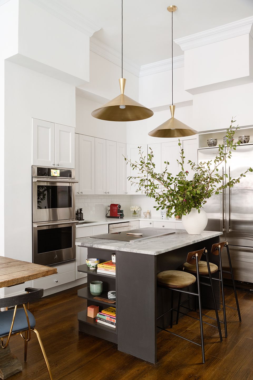 Contemporary kitchen with white cabinetry, brass pendant lights, dark island, and minimalist aesthetic with natural greenery.