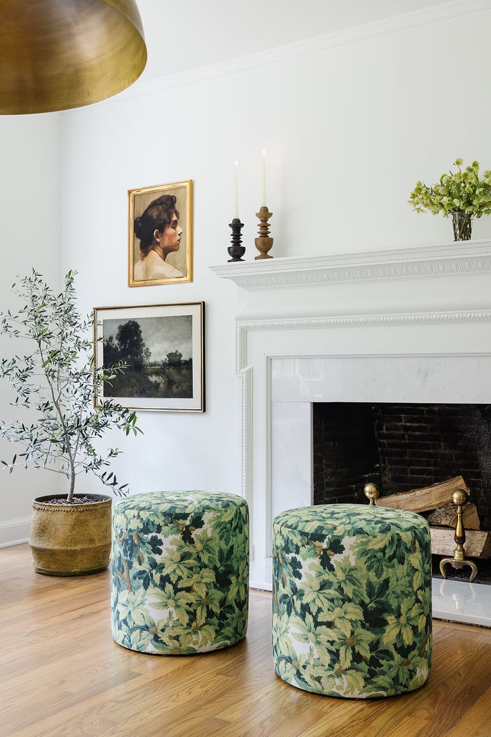 Contemporary fireplace vignette with botanical-printed poufs, brass accents, white mantel styling, and botanical artwork creating elevated, curated feel