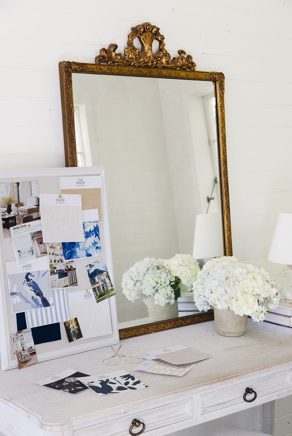 Elegant vanity with ornate gold mirror, white florals, and mood board styling on white desk