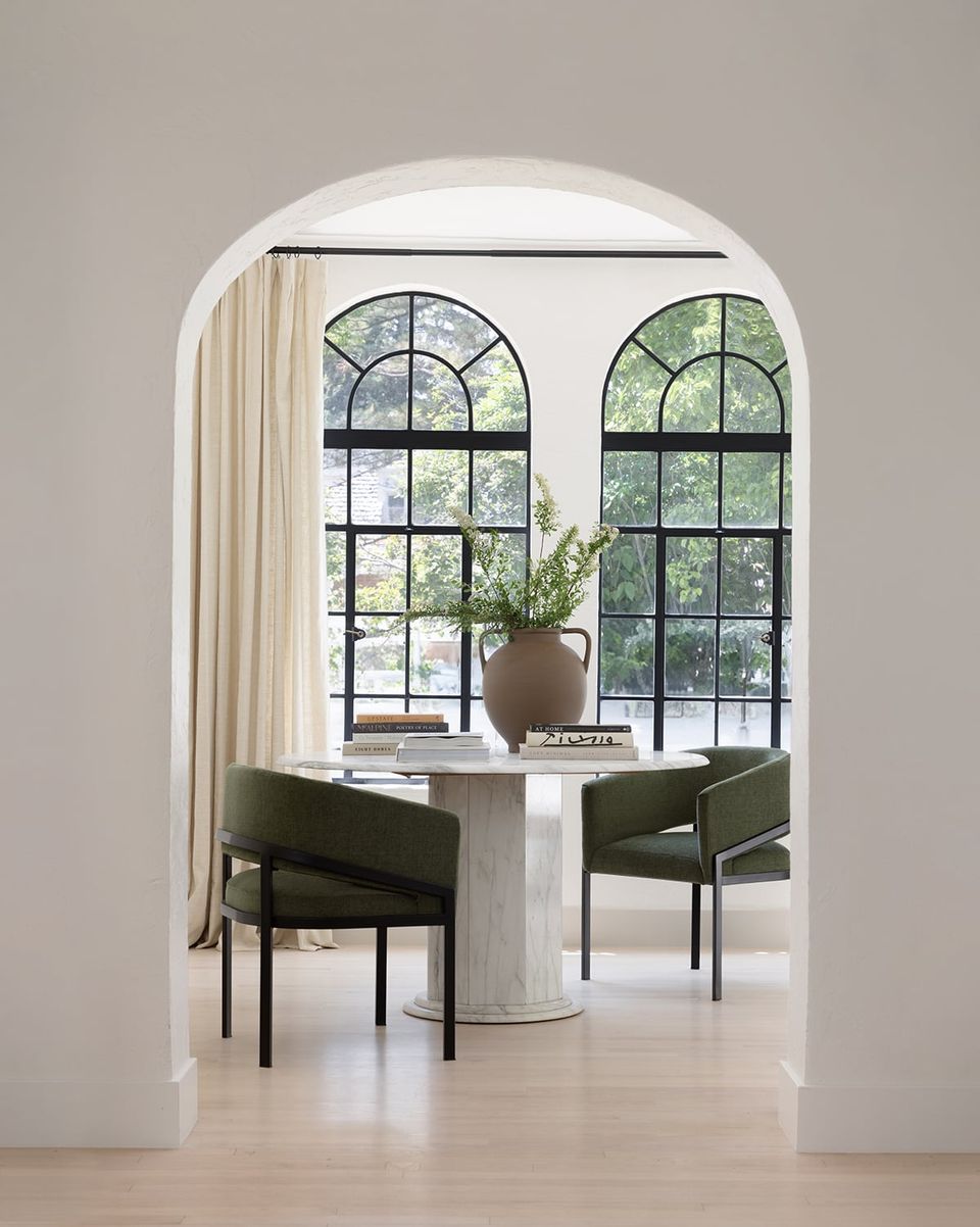 Modern minimalist dining room featuring arched windows, marble pedestal table, and olive green chairs in serene white space