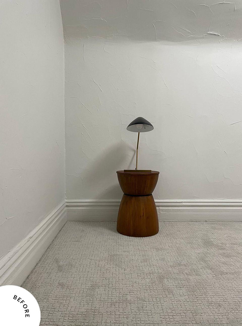 Minimalist before space: bare white walls, wooden pedestal table, and simple black lamp on neutral carpet.