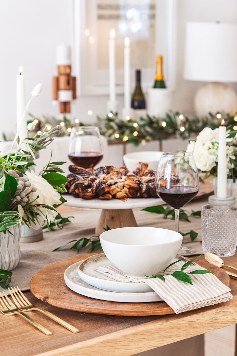 Modern holiday tablescape with natural wood, white ceramics, fresh greenery, and warm candlelight in neutral tones