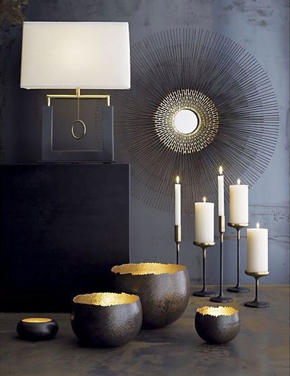 Dark moody vignette with brass sunburst mirror, ceramic bowls, white lamp, and candleholder creating dramatic luxury ambiance.
