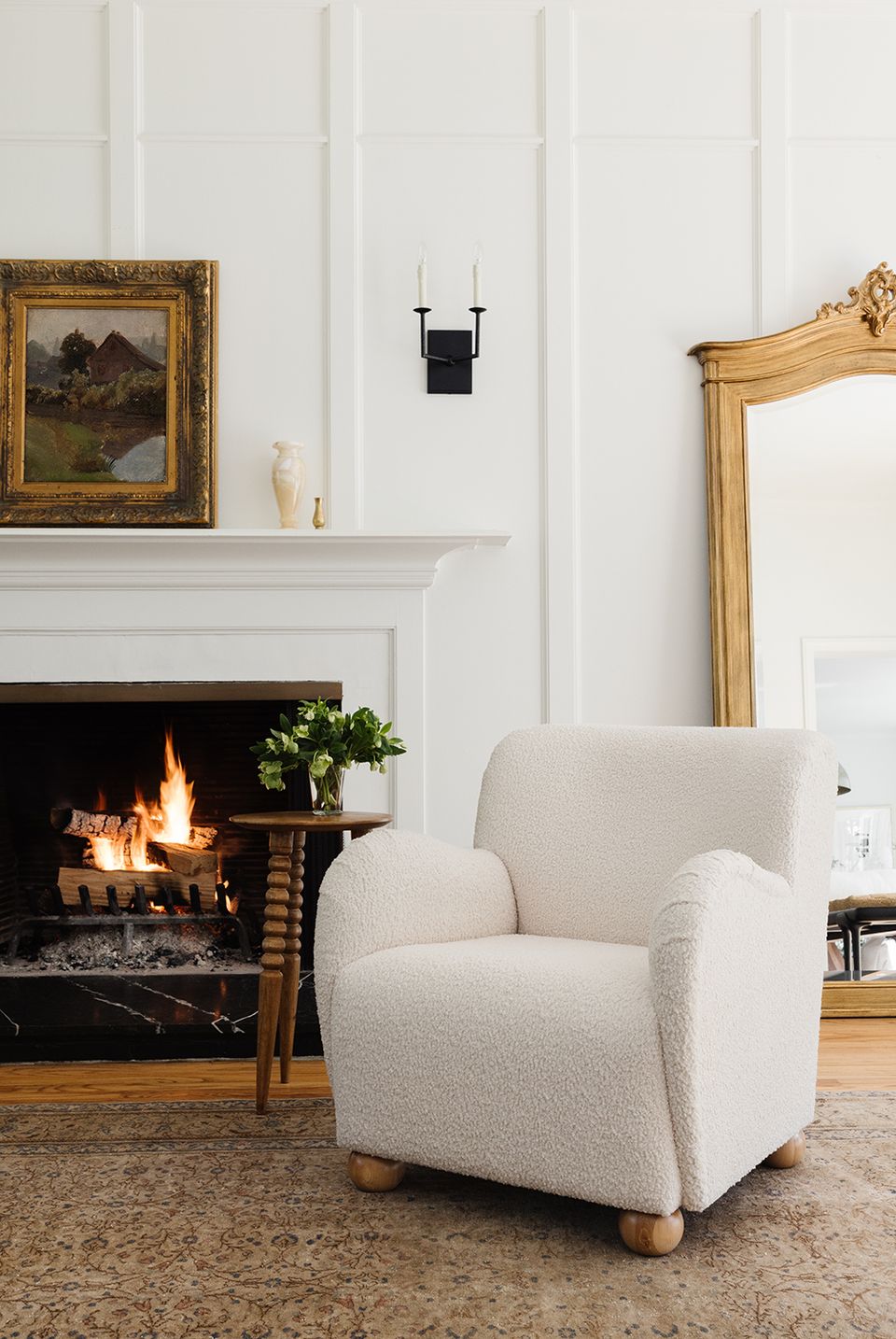 Refined fireplace nook with classic white paneling, cream boucle armchair, glowing hearth, and gold-framed accents blending traditional elegance with modern minimalism.