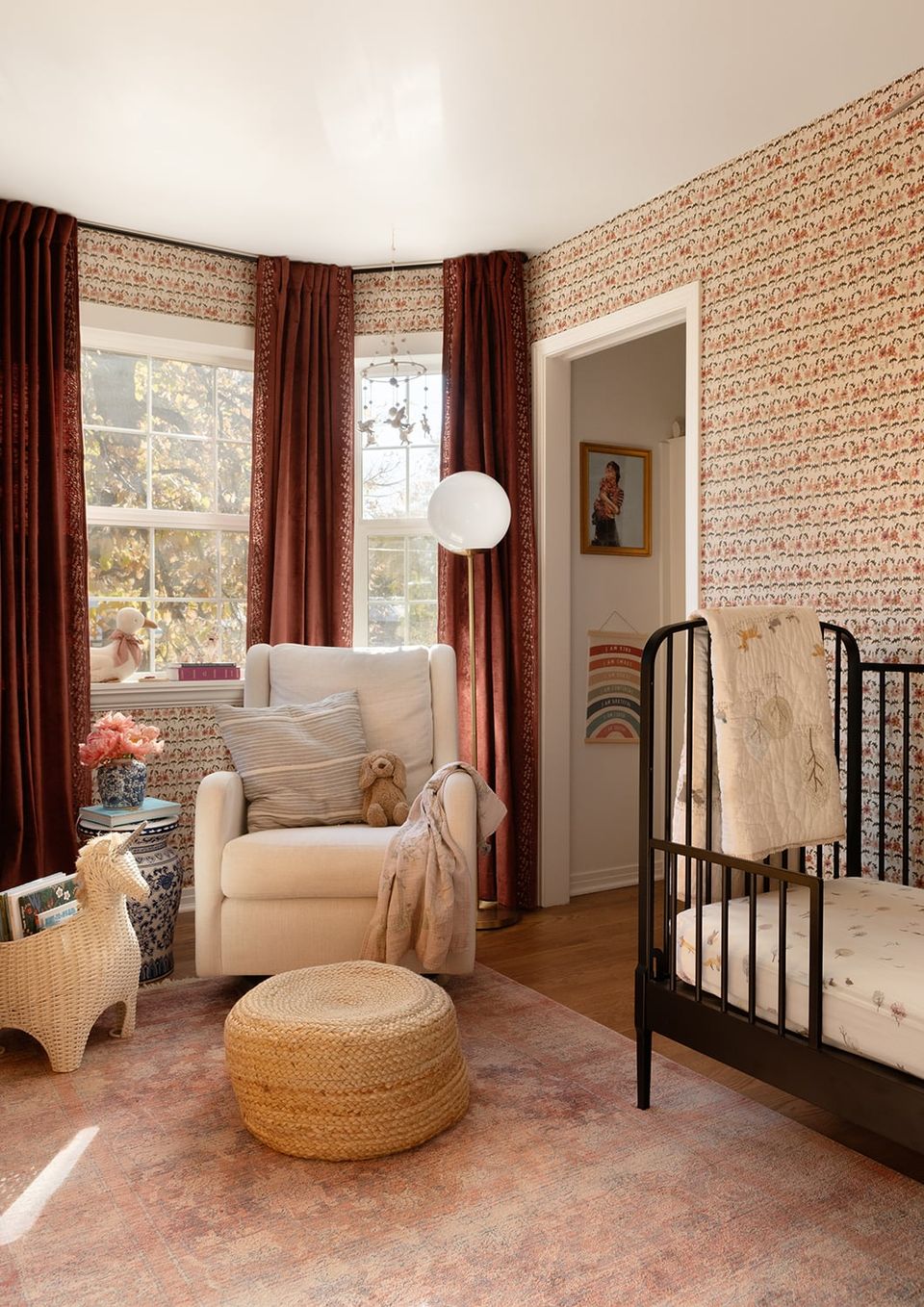 Warm, eclectic nursery with terracotta patterned walls, burgundy curtains, natural wood accents, and vintage-inspired furnishings in cream and tan tones.